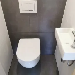 Nowoczesna, mała łazienka z podwieszaną toaletą i umywalką, szare płytki na ścianie i podłodze, biała armatura, minimalistyczny styl.