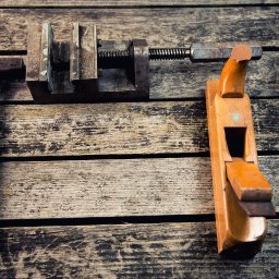 MEBLE NA WYMIAR NV Service - Stare narzędzia stolarskie: młotek z niebieskim trzonkiem, ścisk stolarski i strug na tle desek z widoczną strukturą drewna. Vintage.