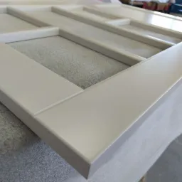 Rama drewniana z podziałami, pomalowana na jasny kolor, leżąca na szarej powierzchni, widok z bliska na narożnik ramy.