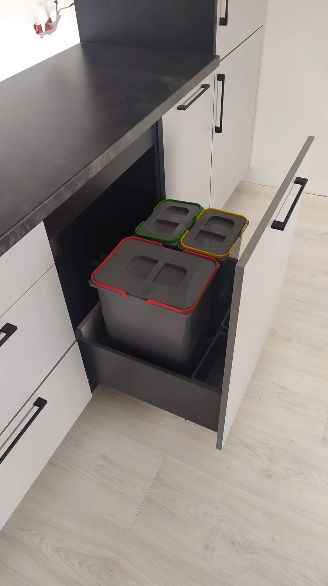 Wysuwany kosz na śmieci z trzema pojemnikami w szafce kuchennej z białymi frontami i czarnymi uchwytami, widok z góry, jasne panele podłogowe.
