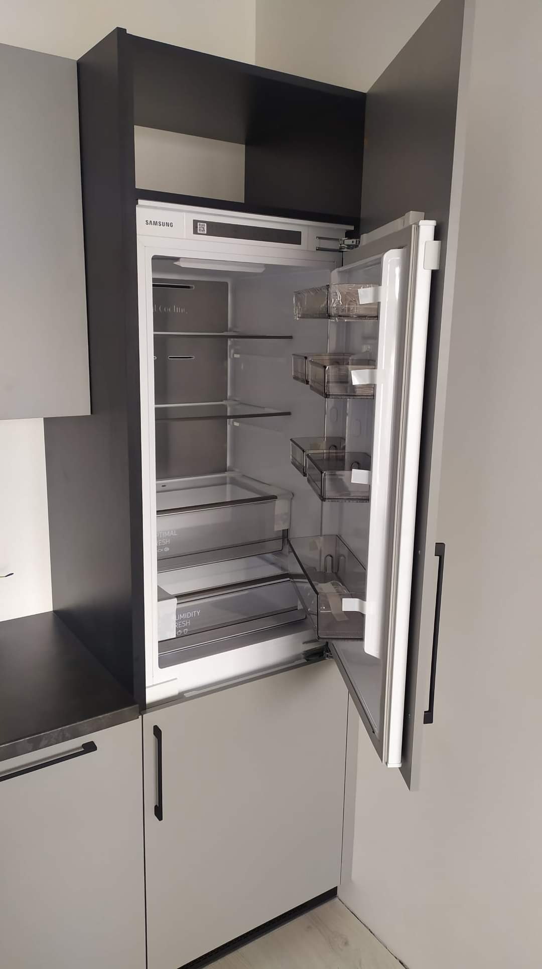 Wbudowana lodówka Samsung w zabudowie kuchennej z szarymi szafkami i czarnymi elementami, drzwi lodówki otwarte, widoczne półki i szuflady.