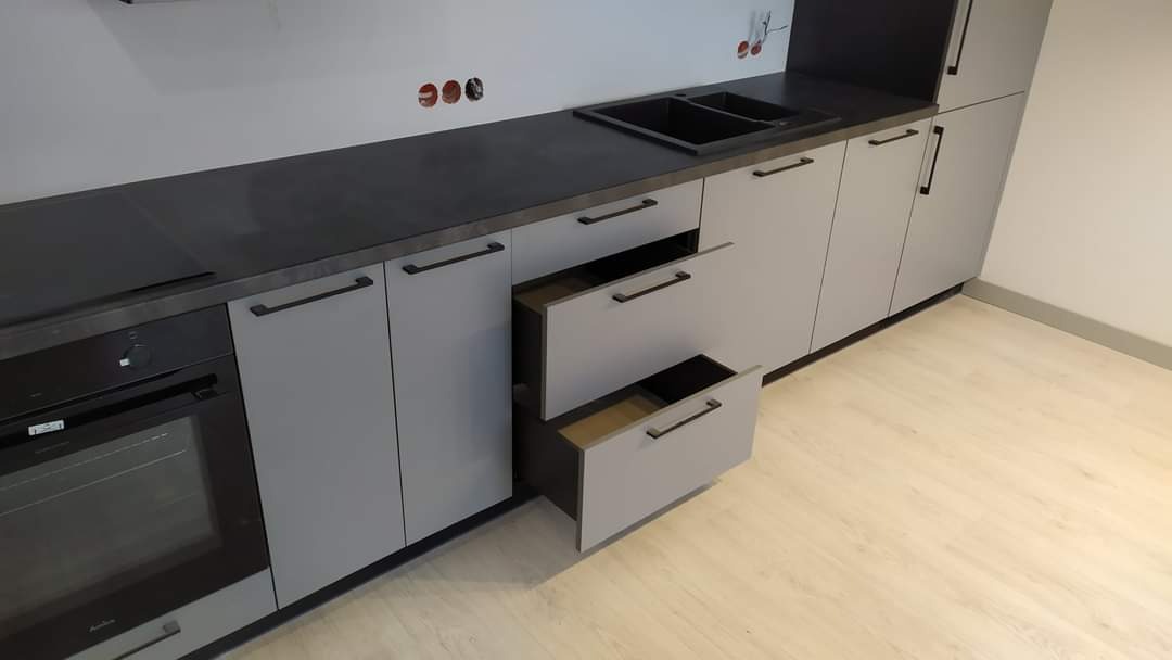 Szara zabudowa kuchenna z ciemnym blatem i otwartymi szufladami na tle jasnej podłogi i ściany z widocznymi puszkami elektrycznymi.