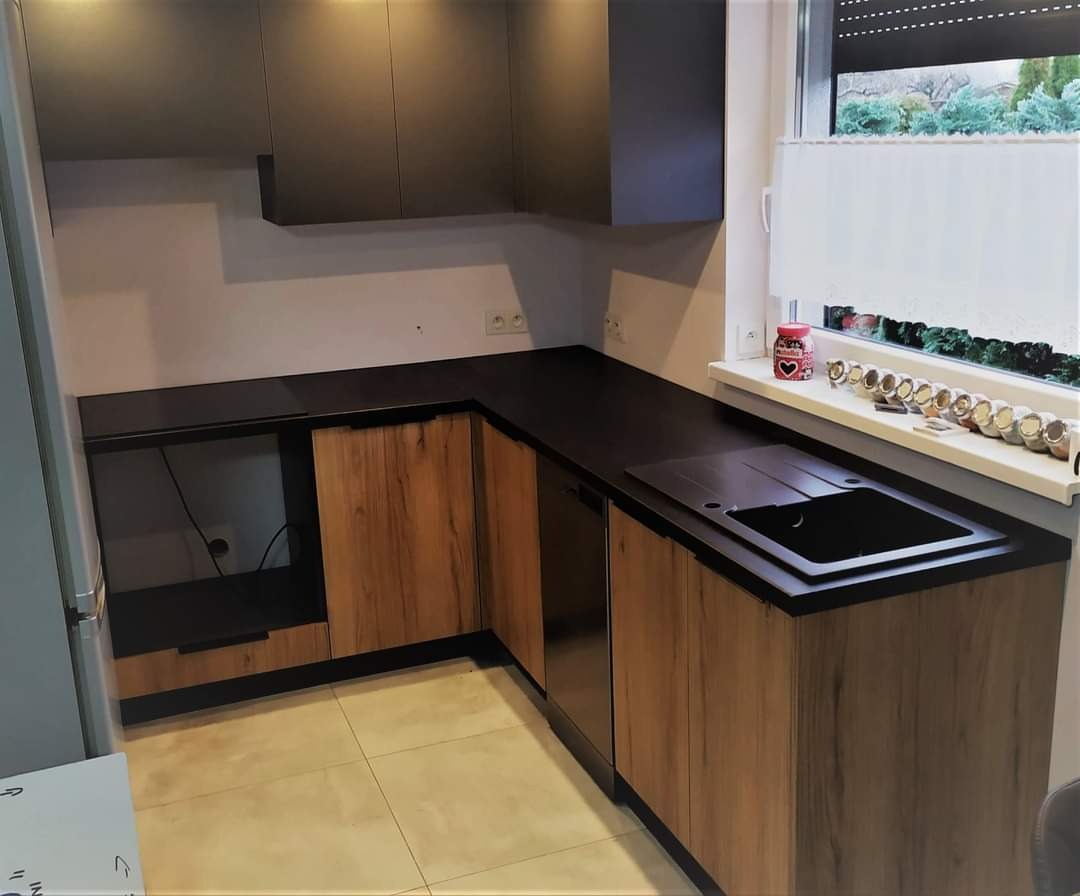 Nowoczesna kuchnia w odcieniach szarości i drewna z czarnym blatem, widok na okno z przyprawami i roletami, neutralne płytki podłogowe.