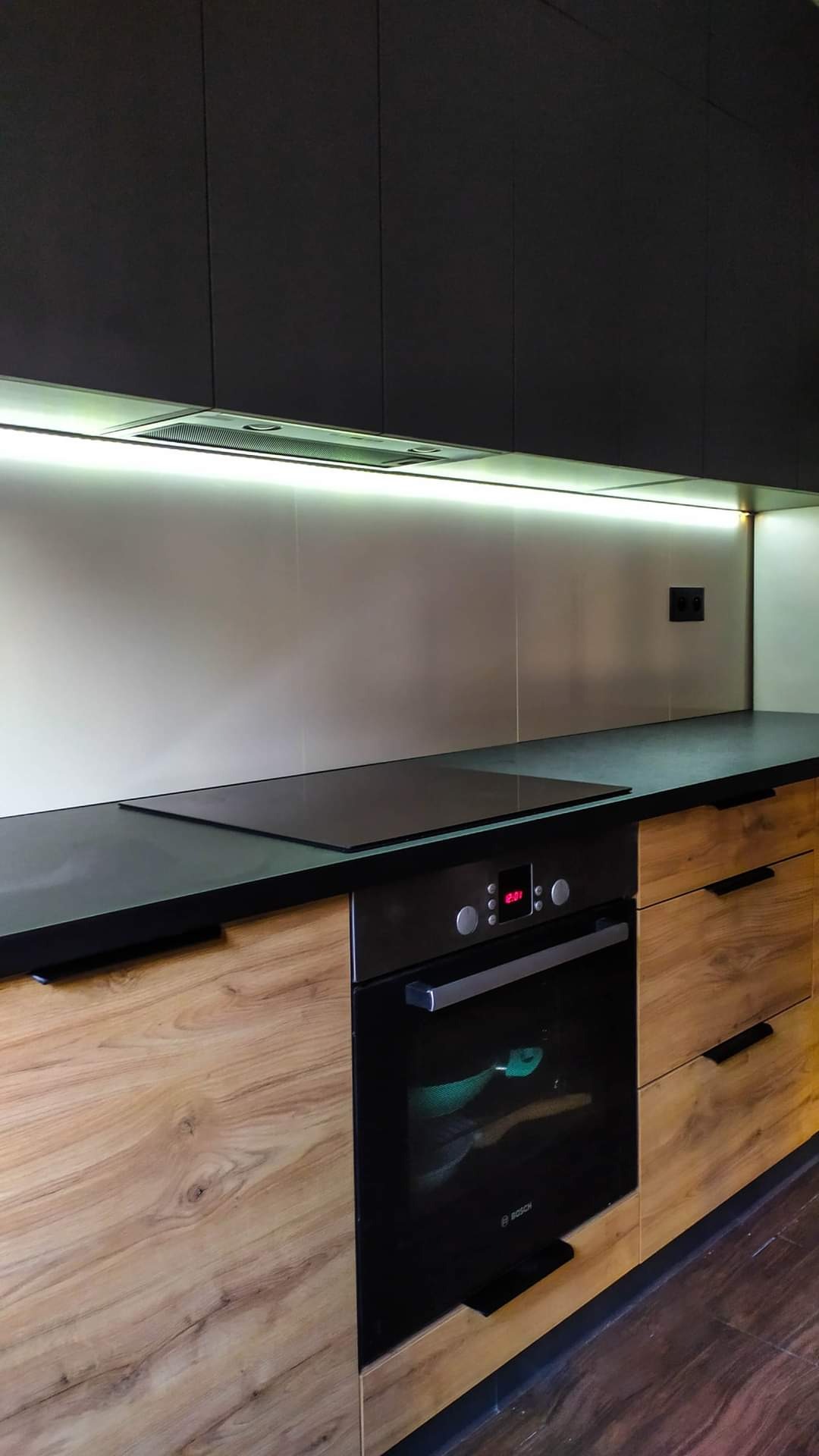 Nowoczesna zabudowa kuchenna z drewnopodobnymi frontami szafek dolnych, czarnymi szafkami górnymi i blatem, płyta indukcyjna i piekarnik Bosch, oświetlenie LED pod szafkami.
