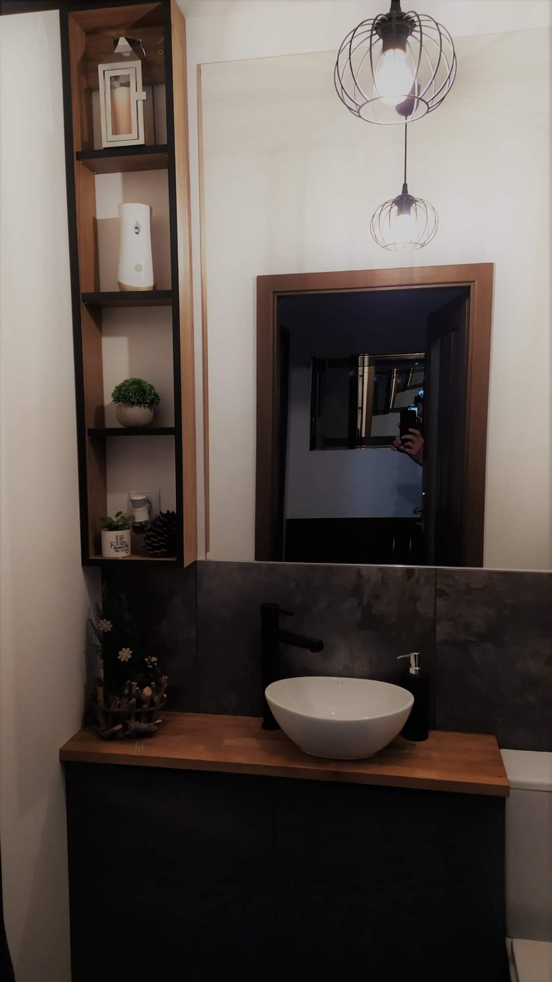 Nowoczesna łazienka z umywalką nablatową, drewnianym blatem, czarną armaturą i szafką w kolorze antracytu, dodatkowo półka z dekoracjami i lustro z drewnianą ramą.