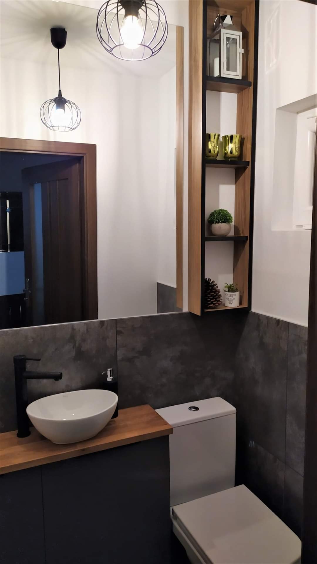 Nowoczesna łazienka z umywalką nablatową, drewnianym blatem, szafką w kolorze grafitowym, wiszącą szafką z drewnianymi półkami i dekoracjami, oraz dwoma lampami wiszącymi w stylu industrialnym.