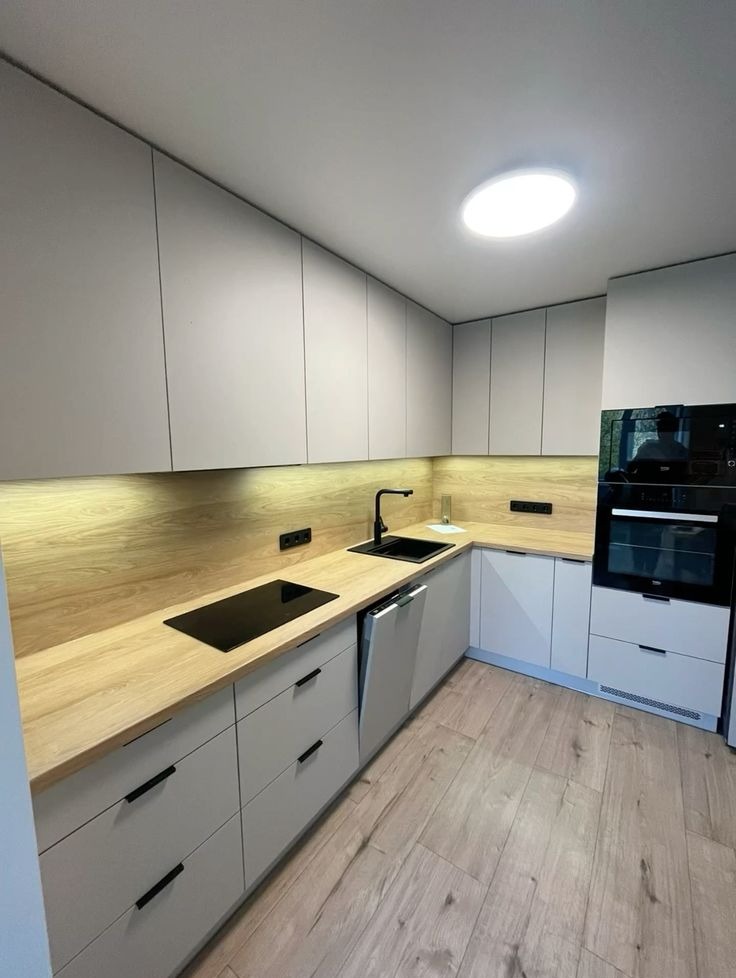 Jasna kuchnia w stylu skandynawskim z drewnianym blatem i frontami w kolorze gołębim. Minimalistyczne uchwyty, płyta indukcyjna i czarny kran dodają elegancji.