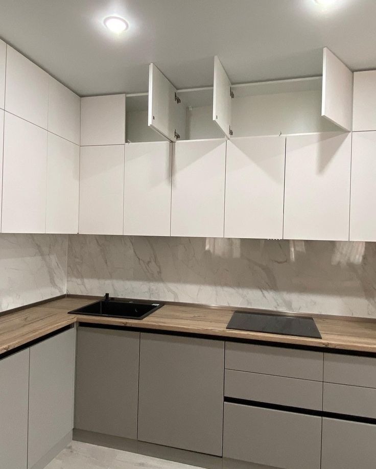 Nowoczesna kuchnia z białymi szafkami górnymi (część otwarta) i szarymi dolnymi, drewniany blat, marmurowa ściana. Minimalistyczny design, oświetlenie sufitowe.