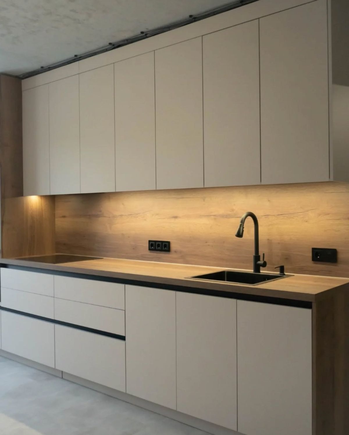 Nowoczesna kuchnia z beżowymi frontami szafek, drewnianym blatem i czarną armaturą. Minimalistyczny design, oświetlenie LED pod szafkami górnymi. Widoczne gniazdka elektryczne.