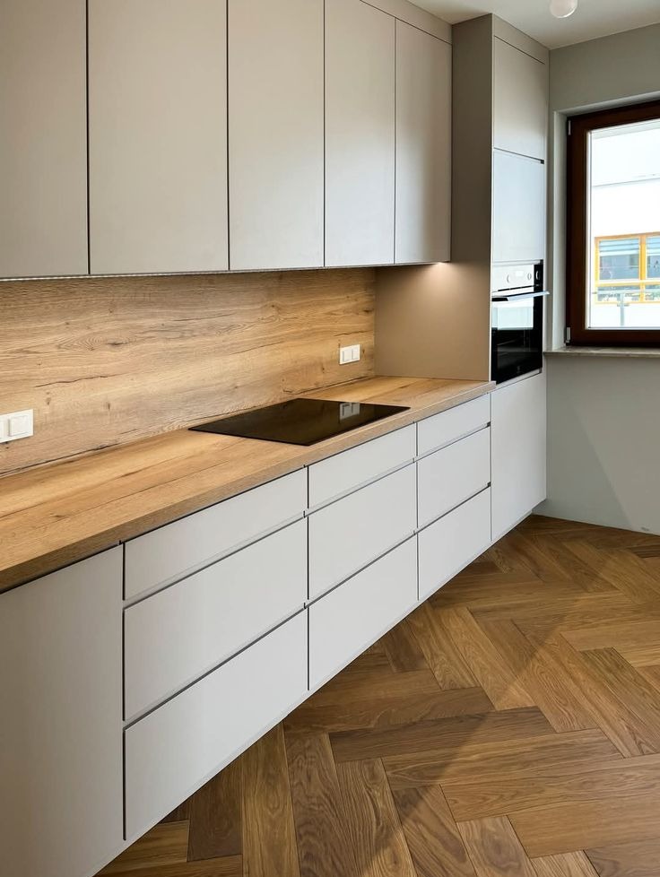Jasnoszara kuchnia z drewnianym blatem i panelem, widok na płytę indukcyjną i parkiet ułożony w jodełkę. Minimalistyczny design, nowoczesne oświetlenie podszafkowe.