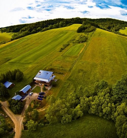 Widok z lotu ptaka na ośrodek wypoczynkowy z niebieskimi dachami, położony na zboczu zielonego wzgórza, otoczony lasem i polami.