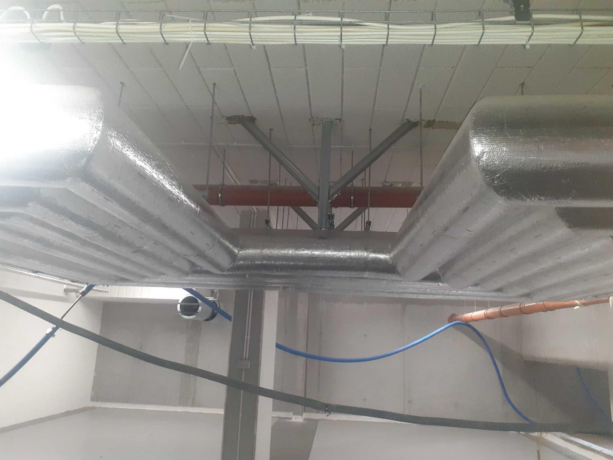 Widok z dołu na instalację wentylacyjną z izolacją termiczną, rury instalacyjne w kolorze pomarańczowym i niebieskim, oraz kable podwieszone pod sufitem w budynku w stanie surowym.