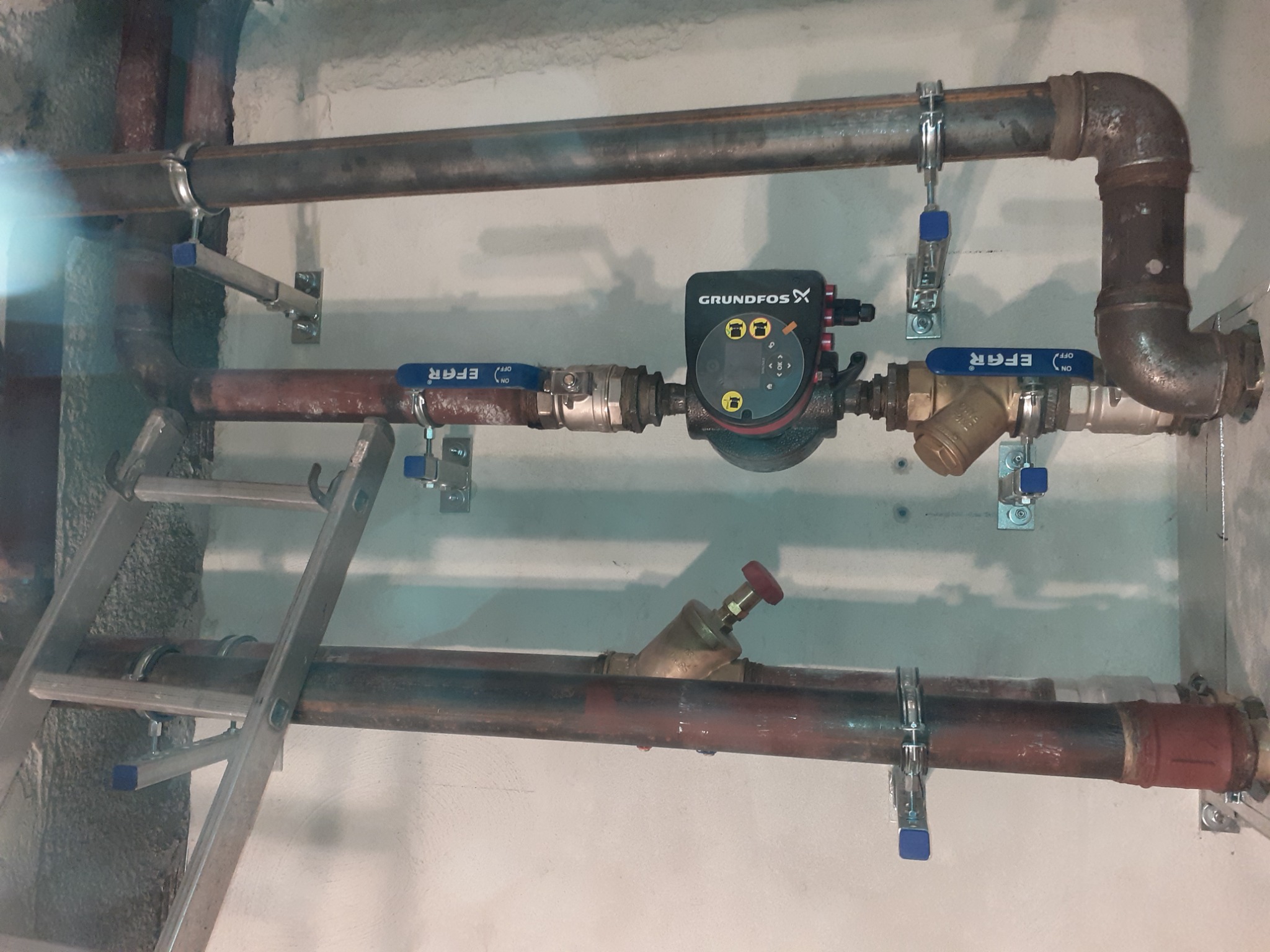 Fragment instalacji hydraulicznej z pompą obiegową Grundfos, zaworami EFAR i filtrem skośnym, rury miedziane mocowane do ściany za pomocą metalowych obejm, w tle drabina.