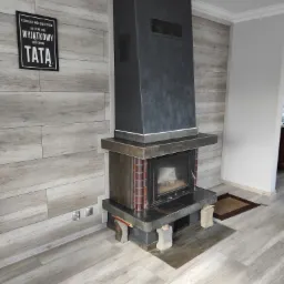Nowoczesny kominek z metalową obudową i ceglanymi akcentami, umieszczony na tle ściany wyłożonej panelami imitującymi drewno. Na ścianie wisi czarna tabliczka z napisem 'TATA'.