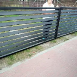 Czarna, metalowa brama z poziomych profili, widoczna z boku, z osobą w tle i fragmentem samochodu po prawej stronie, na tle trawnika i kostki brukowej.