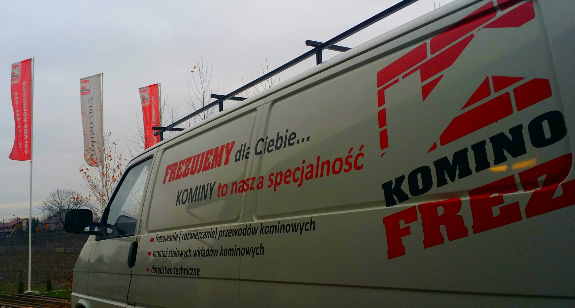 Biały samochód dostawczy z czerwono-czarnym logo firmy oferującej frezowanie przewodów kominowych i montaż wkładów stalowych, na tle trzech flag reklamowych z logo firmy.