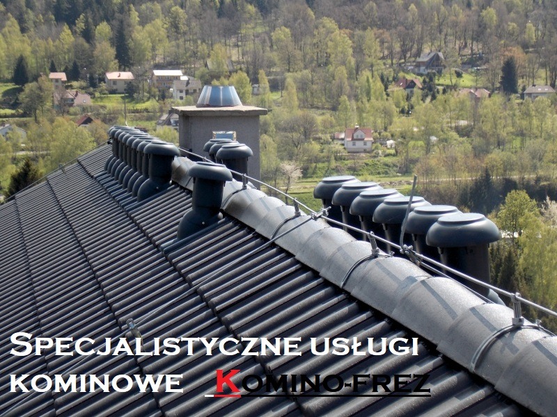 Rząd czarnych nasad kominowych wentylacyjnych na ciemnoszarym dachu z dachówki ceramicznej, z widokiem na pagórkowaty krajobraz z zabudowaniami w tle.