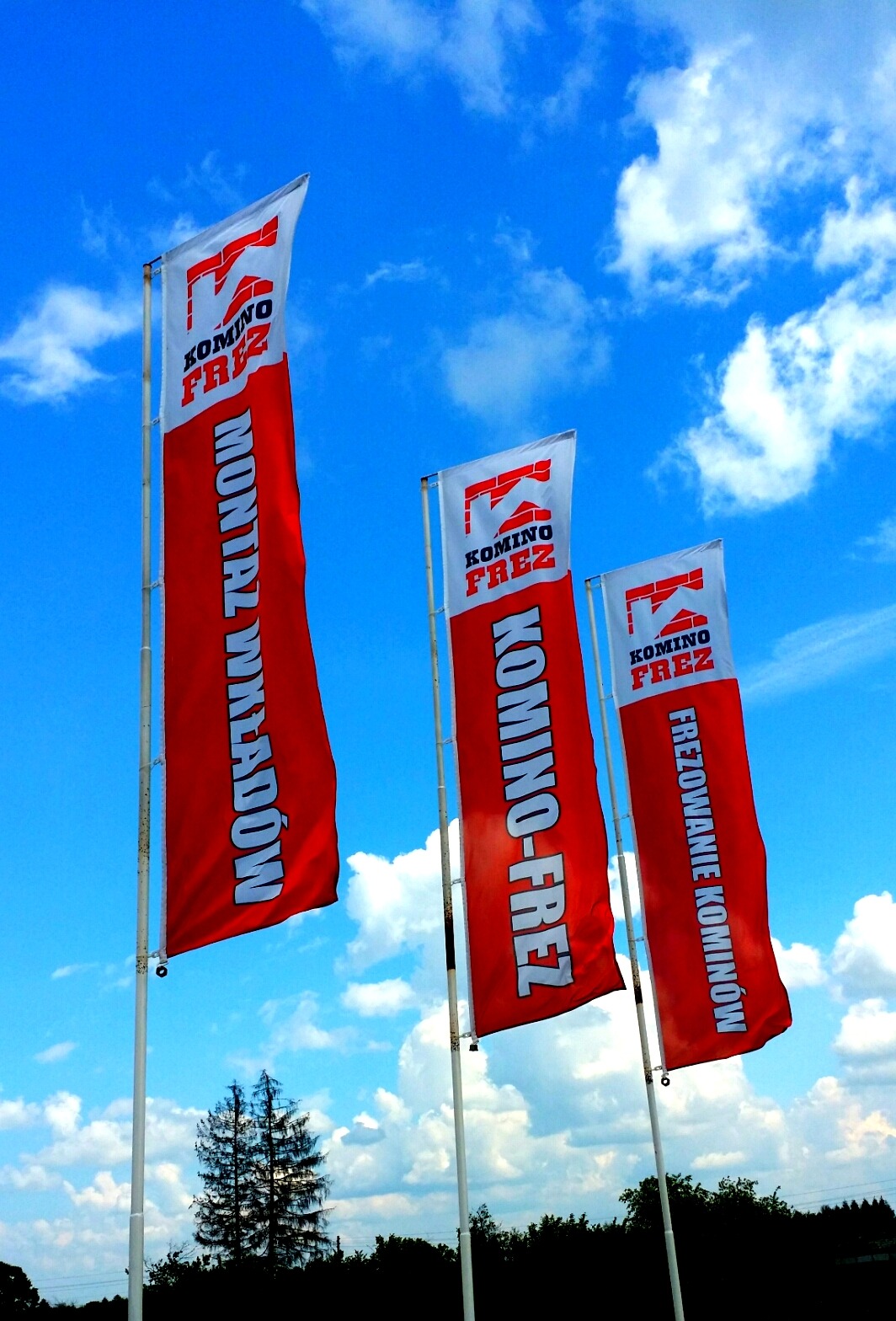 Trzy czerwone flagi reklamowe z logo i napisami 'Montaż wkładów', 'Komino-Frez' i 'Frezowanie kominów' na tle błękitnego nieba z chmurami.