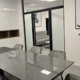 Nowoczesna sala konferencyjna z marmurowym stołem, szarymi krzesłami i rośliną w czarnej donicy, widok na poczekalnię przez przeszklone drzwi.