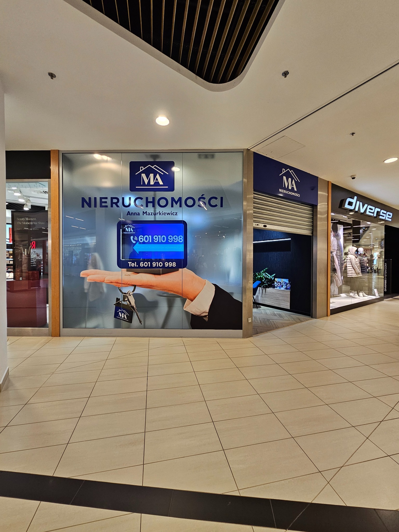 Wnętrze galerii handlowej z witryną biura nieruchomości 'MA Nieruchomości' z grafiką dłoni trzymającej klucze i numerem telefonu. Obok widoczne inne sklepy.
