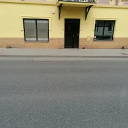 Żółta fasada budynku z ciemnymi drzwiami i oknami, wykończona płytkami imitującymi cegłę w dolnej części, widok z poziomu ulicy.