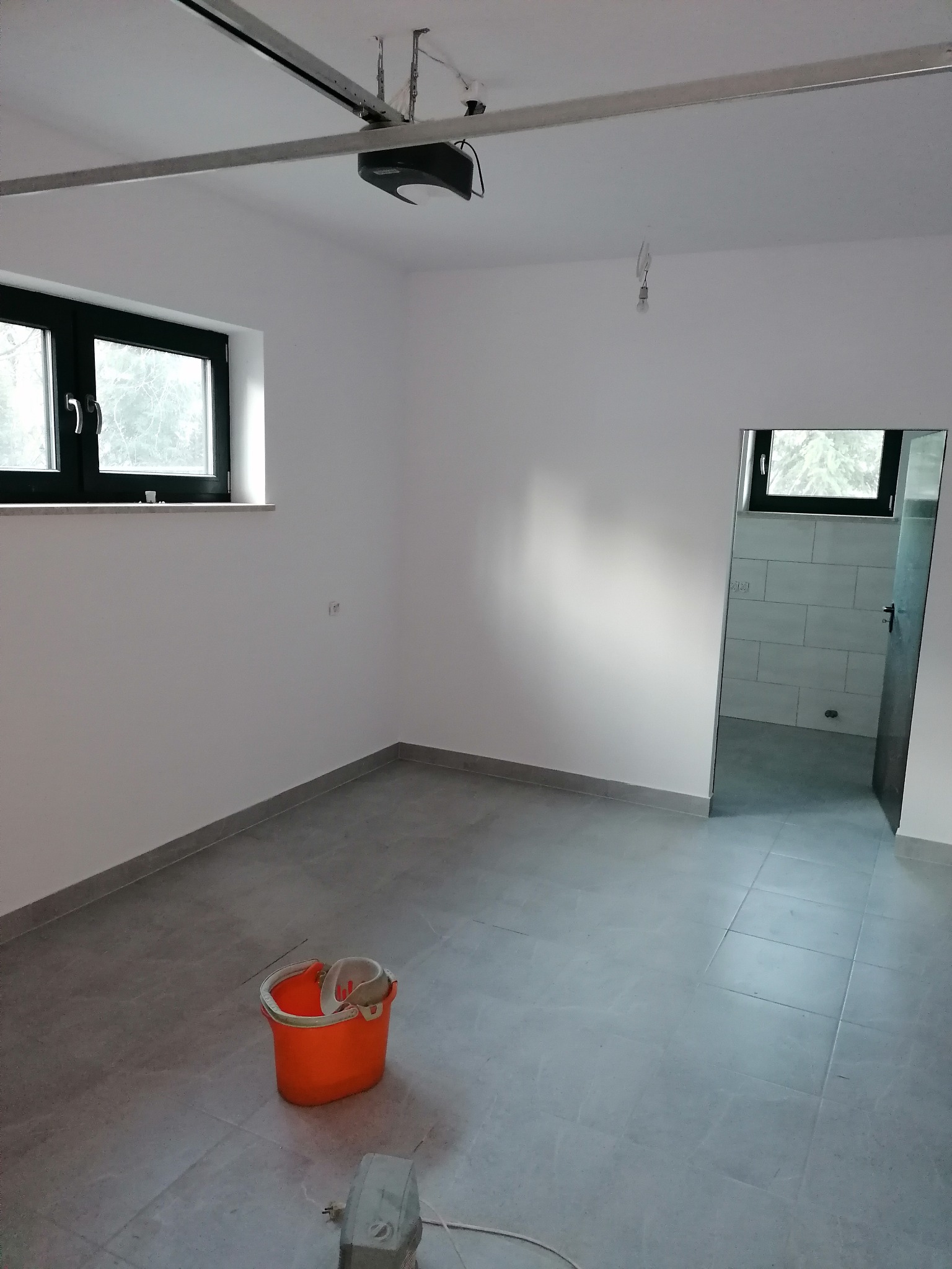 Wnętrze po remoncie z szarymi płytkami podłogowymi, widoczne okno z czarną ramą, pomarańczowe wiadro i odkurzacz na podłodze, oraz odbicie łazienki w lustrze.