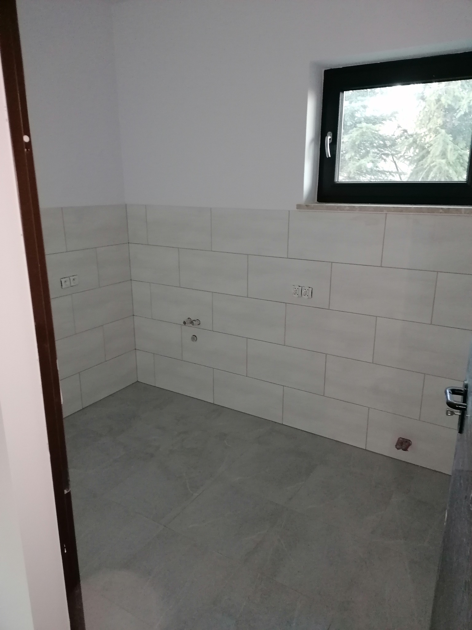 Wykończona łazienka w trakcie remontu z szarymi płytkami podłogowymi i białymi płytkami ściennymi, widoczne przyłącza wodne i okno z widokiem na drzewa.