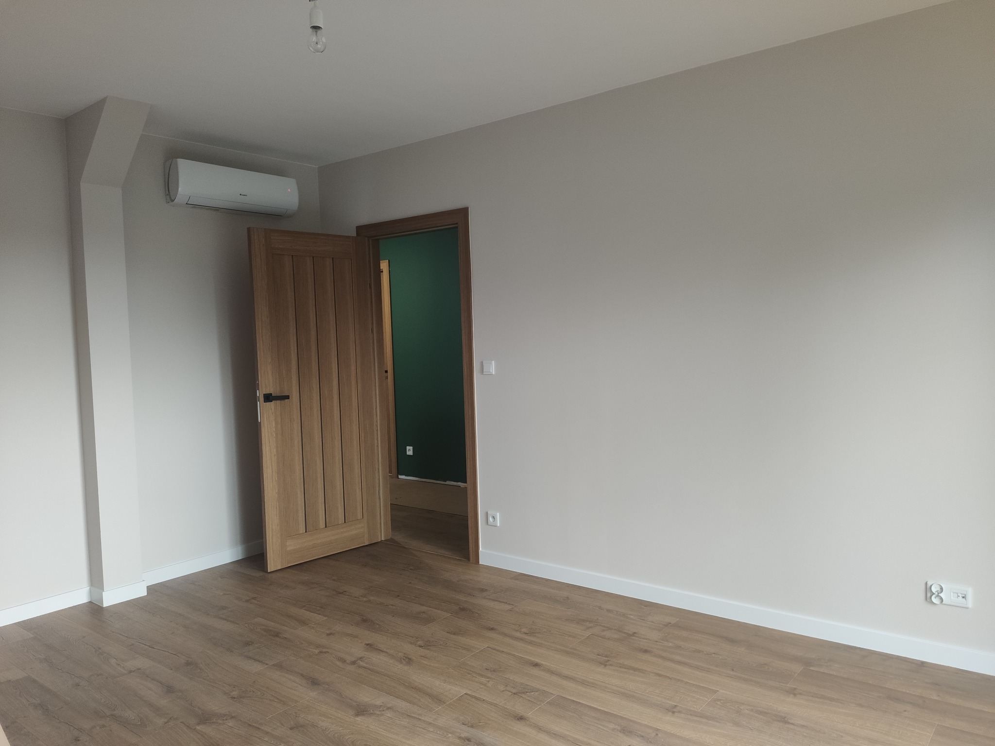Jasne wnętrze po remoncie: drewnopodobne panele, szara ściana z klimatyzatorem, drewniane drzwi z czarną klamką otwarte do pokoju z zieloną ścianą.