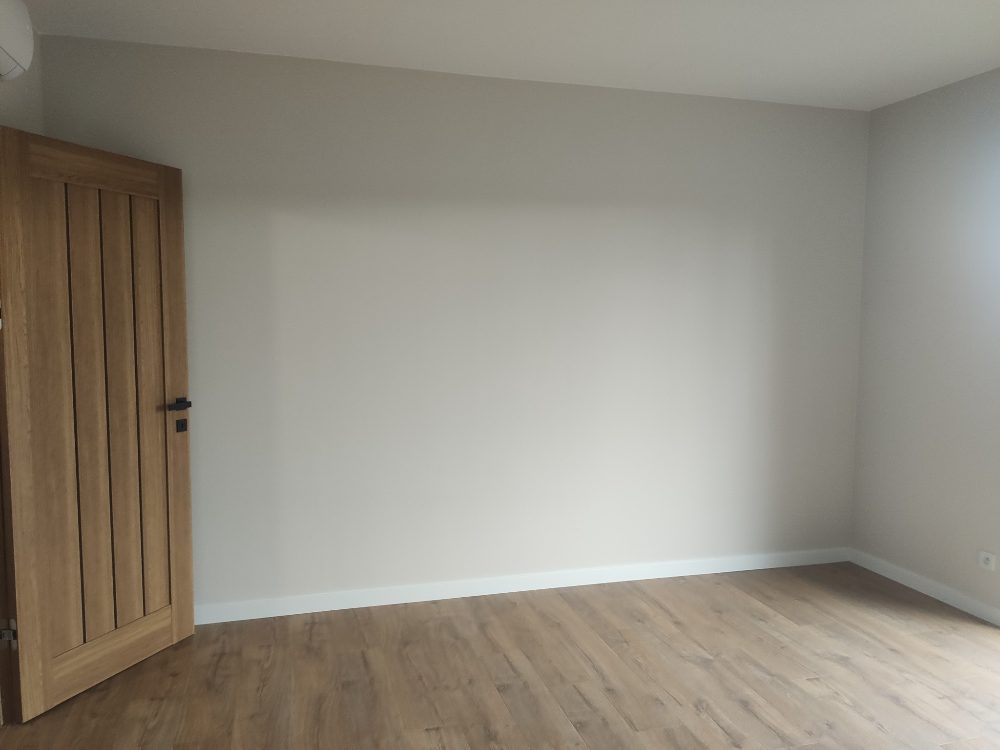 Jasne, puste pomieszczenie z panelami podłogowymi i drewnianymi drzwiami z czarną klamką po lewej stronie.