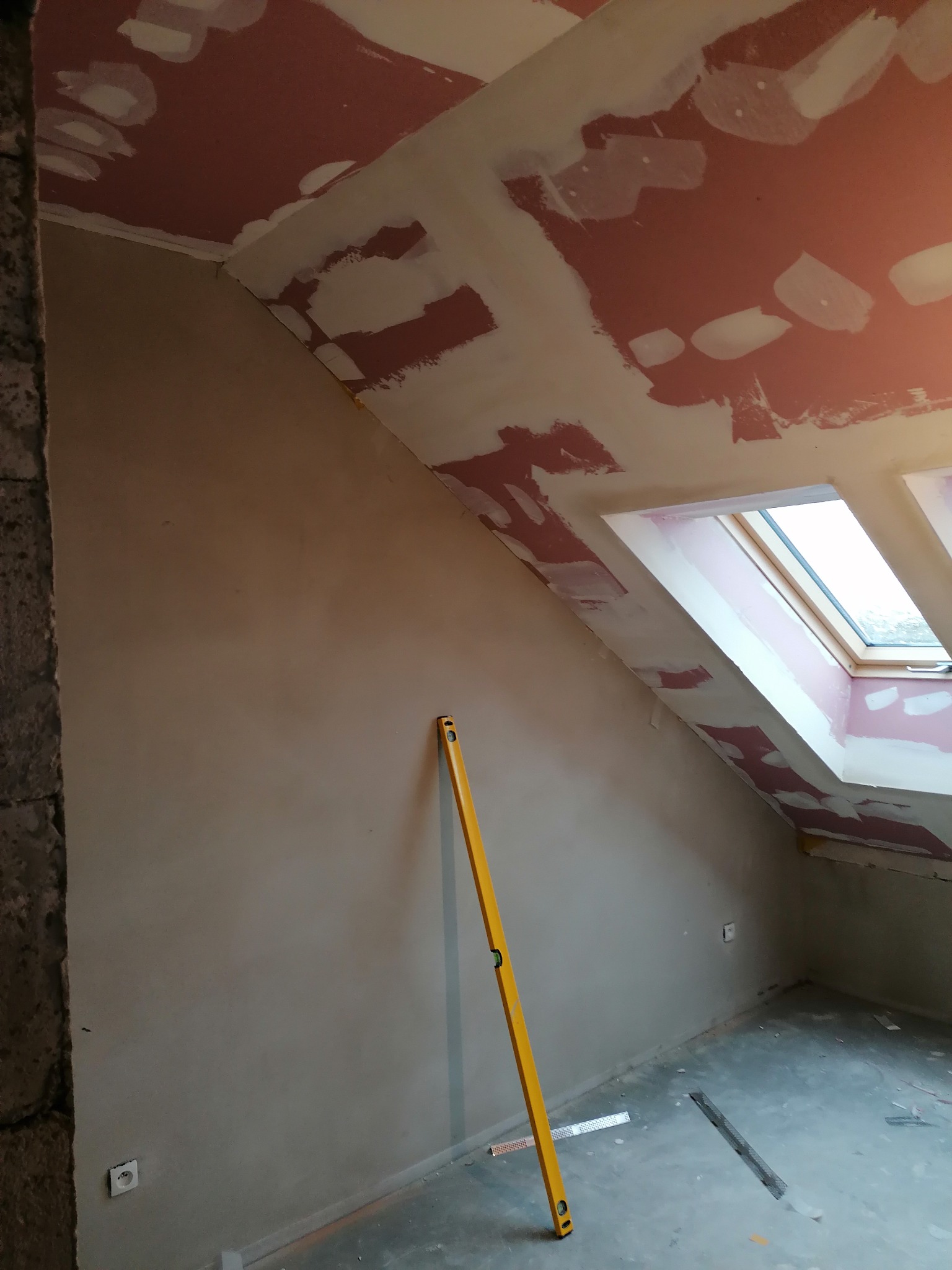 Poddasze w trakcie prac wykończeniowych: ściany i skośny sufit z oknami dachowymi pokryte warstwą szpachli, widoczna żółta poziomica przy ścianie, fragment surowego muru po lewej stronie.