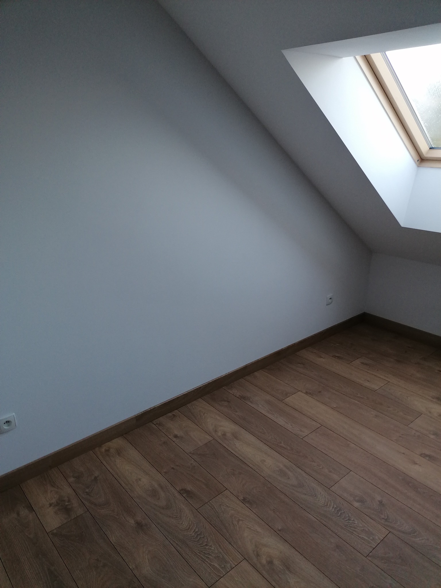 Poddasze po remoncie: Ułożone panele podłogowe w odcieniach brązu, białe ściany i okno dachowe w drewnianej ramie.