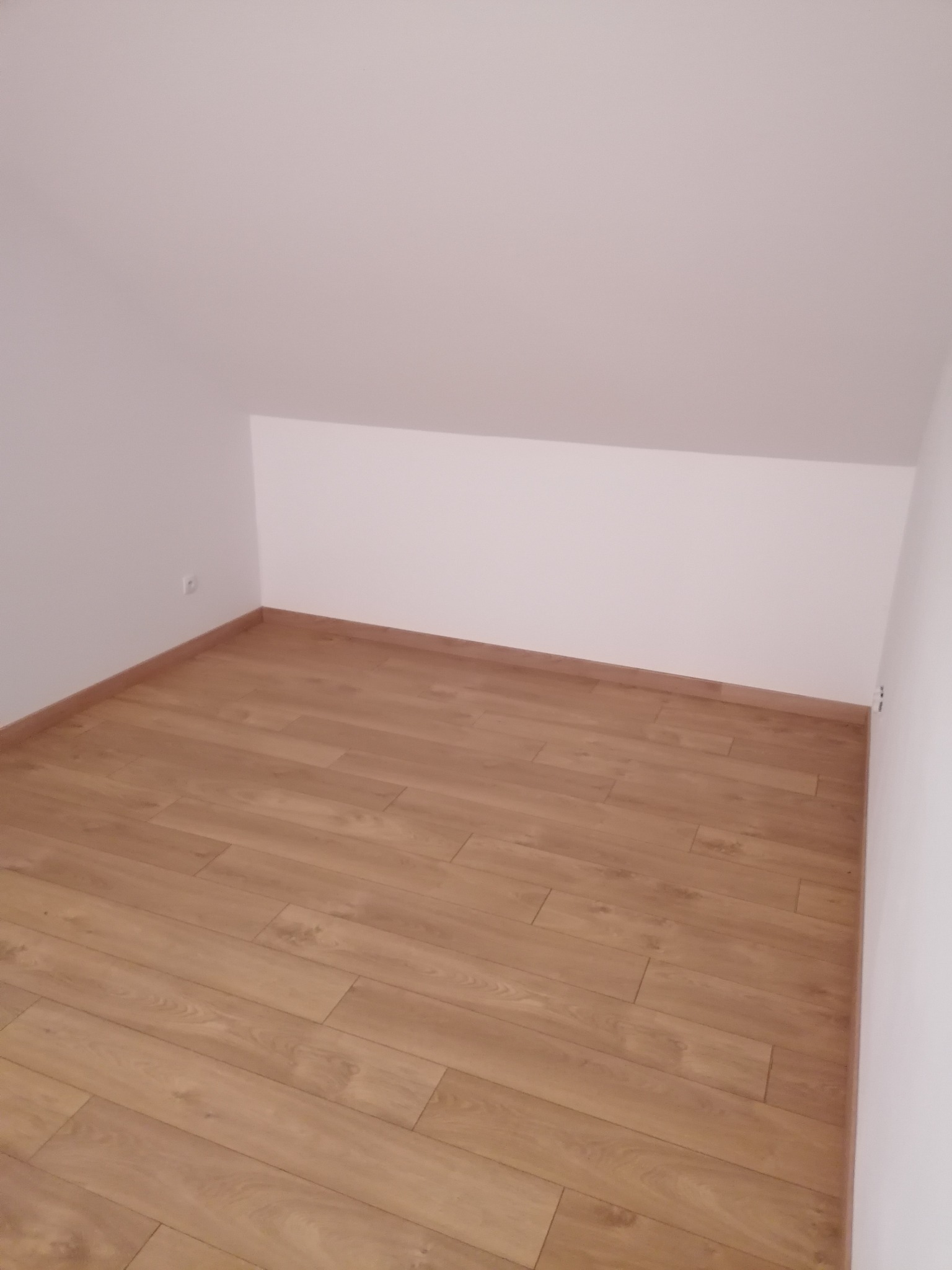 Poddasze po remoncie z ułożonymi panelami podłogowymi w odcieniach naturalnego drewna, widoczna ściana ze skosem i gniazdko elektryczne.