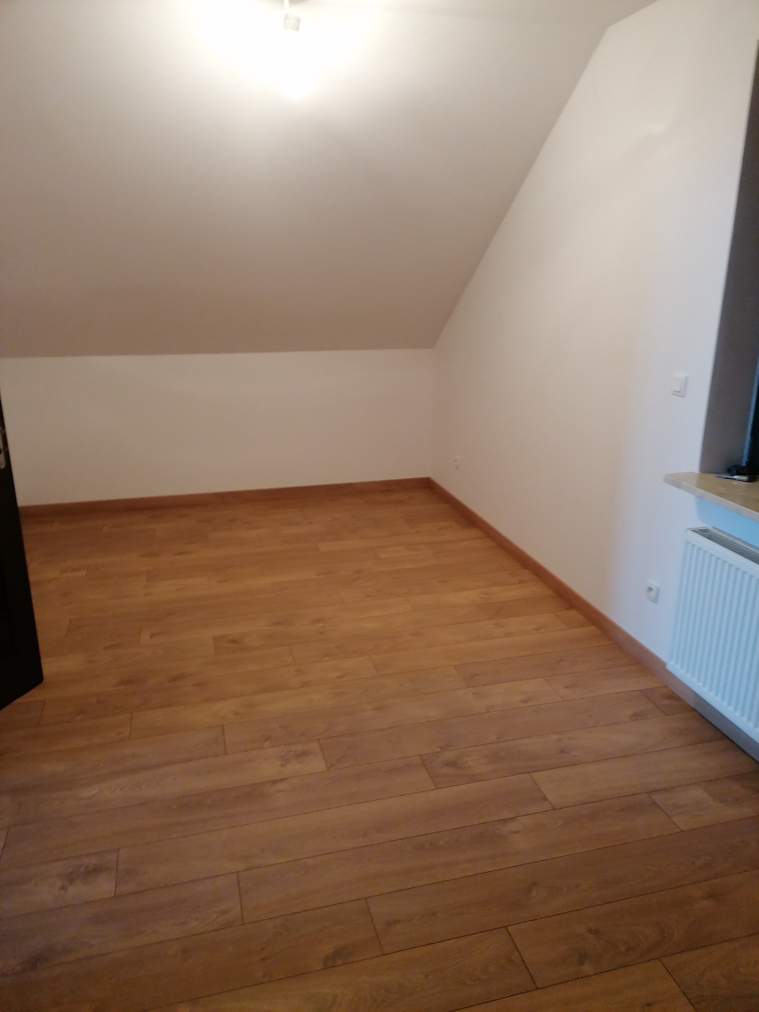 Poddasze z ułożonymi panelami podłogowymi w odcieniach brązu, widoczny grzejnik pod oknem i skosy ścian.