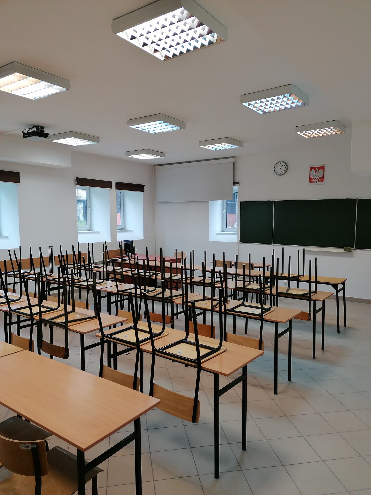 Wnętrze jasnej klasy szkolnej z rzędami pustych ławek, krzesła odwrócone na blatach, białe ściany, okna z roletami, sufit z kwadratowymi lampami, tablica i godło Polski.