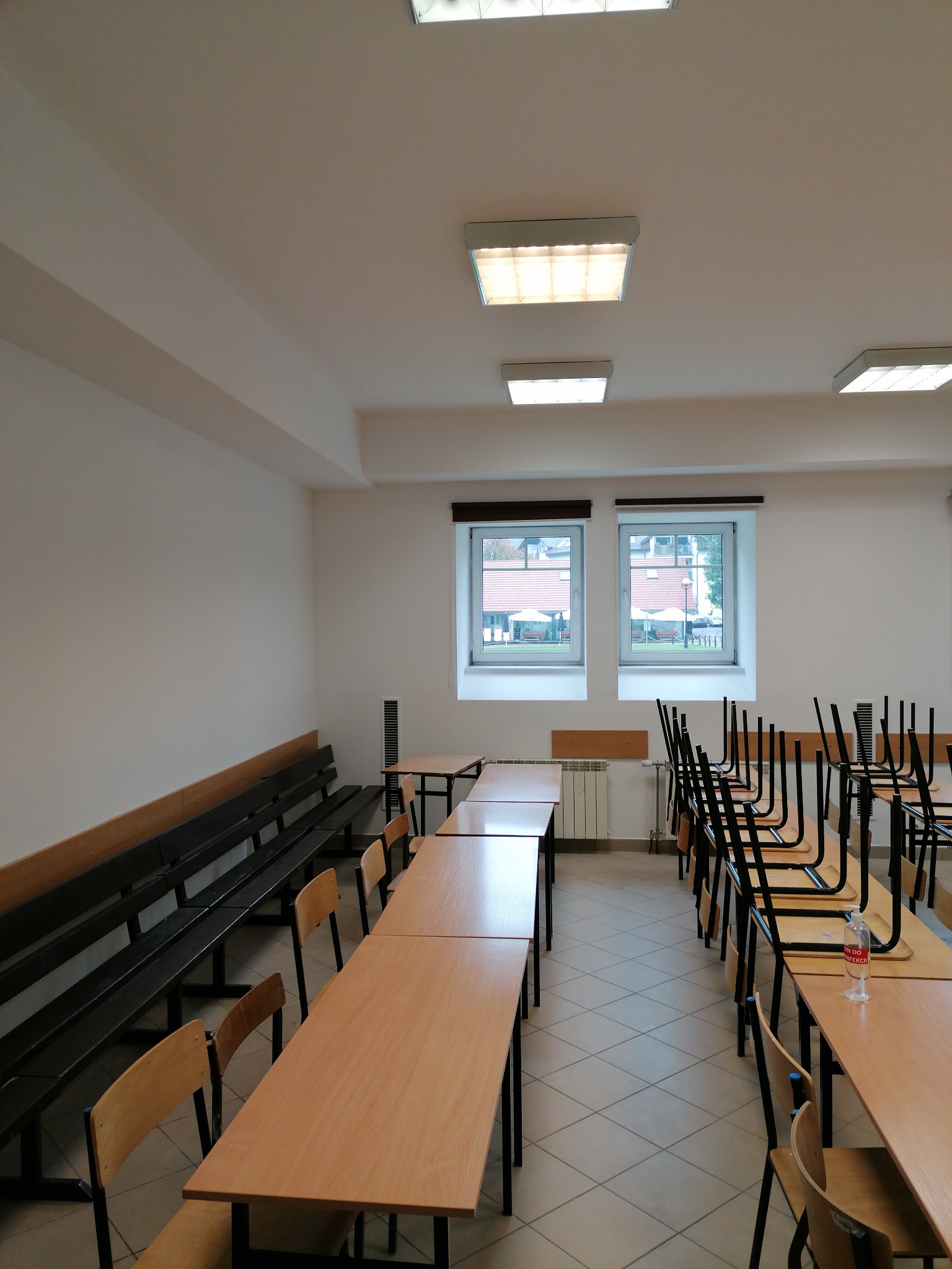 Wnętrze pustej sali z rzędami stolików i krzeseł, część krzeseł odwrócona na stolikach, widoczne okna z widokiem na zewnątrz, białe ściany i sufit z lampami.