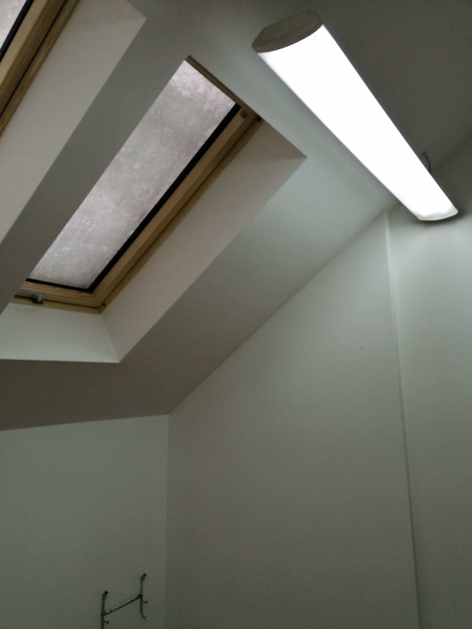 Poddasze z oknem dachowym w drewnianej ramie i podłużną lampą LED, ściany pomalowane na biało, widoczny fragment wieszaka na ręczniki.