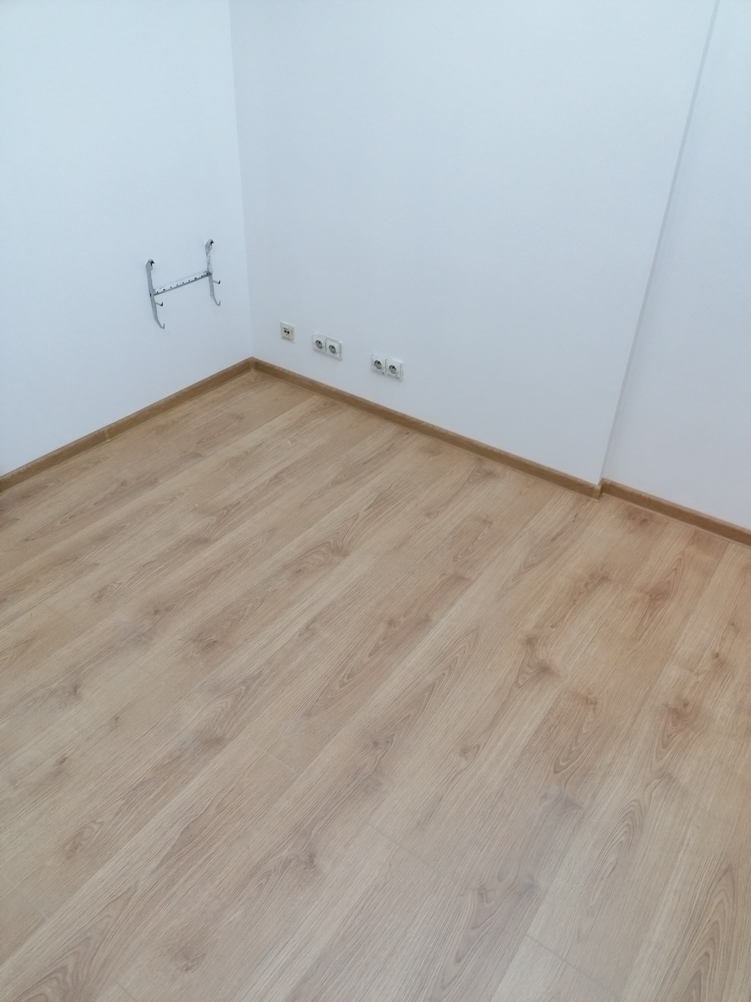 Pokój po ułożeniu paneli podłogowych w odcieniach jasnego drewna, z widocznymi listwami przypodłogowymi, gniazdkami elektrycznymi i nietypowym metalowym wieszakiem na ścianie.