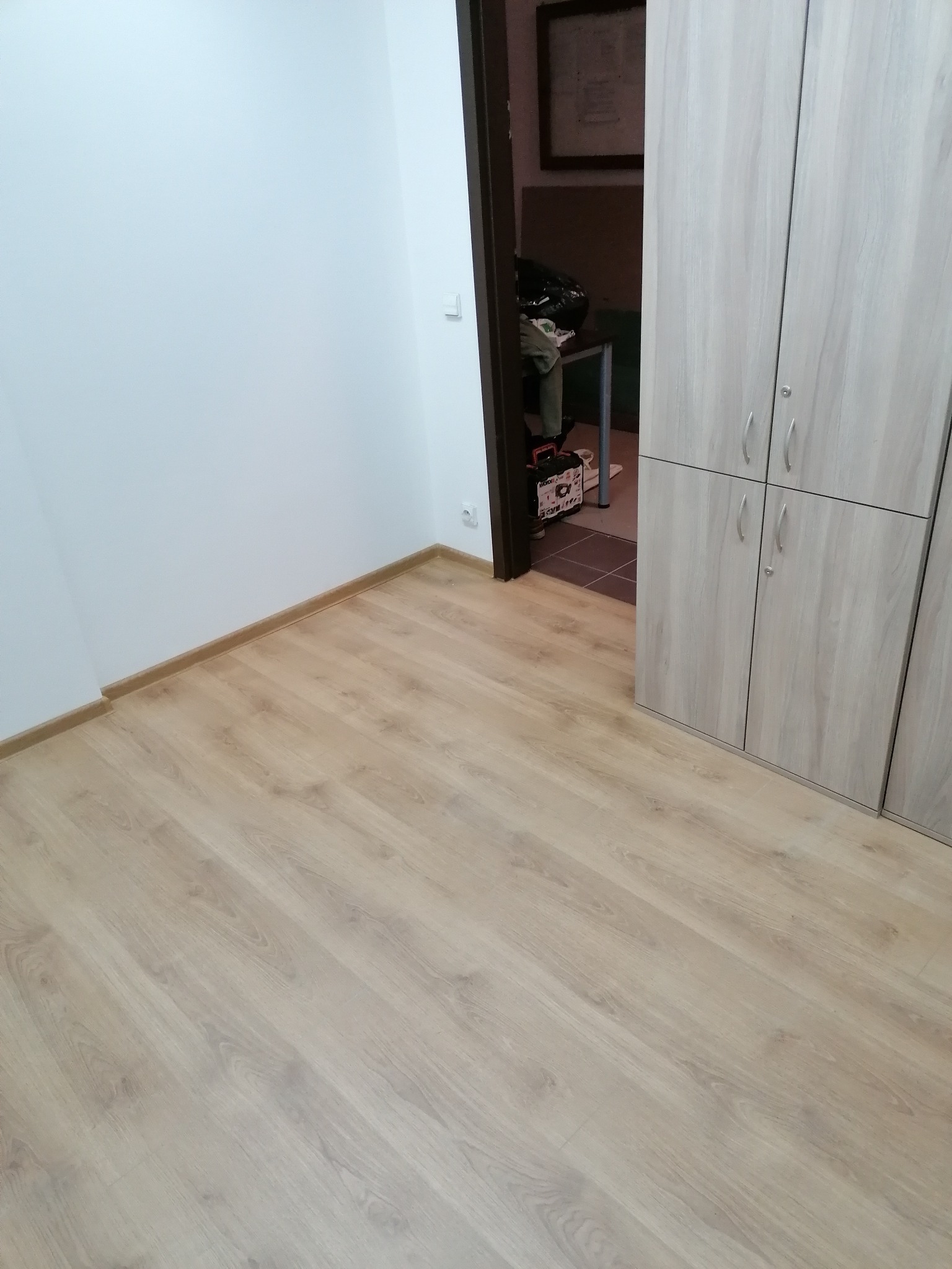 Jasne panele podłogowe ułożone w nowym pomieszczeniu z białymi ścianami i brązowymi listwami przypodłogowymi, widoczna szafa z jasnego drewna i otwarte drzwi do kolejnego pomieszczenia.