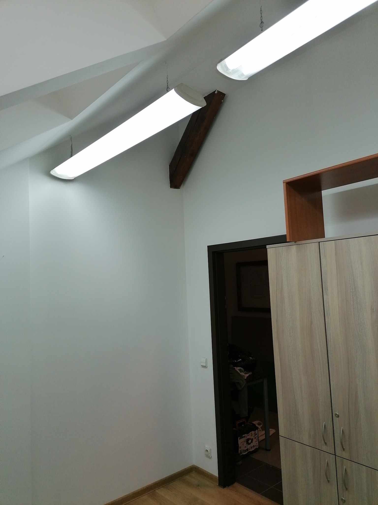 Wnętrze biura po remoncie, widoczne lampy wiszące na łańcuchach, drewniana belka konstrukcyjna, pomalowane ściany, drzwi wejściowe oraz szafa biurowa.