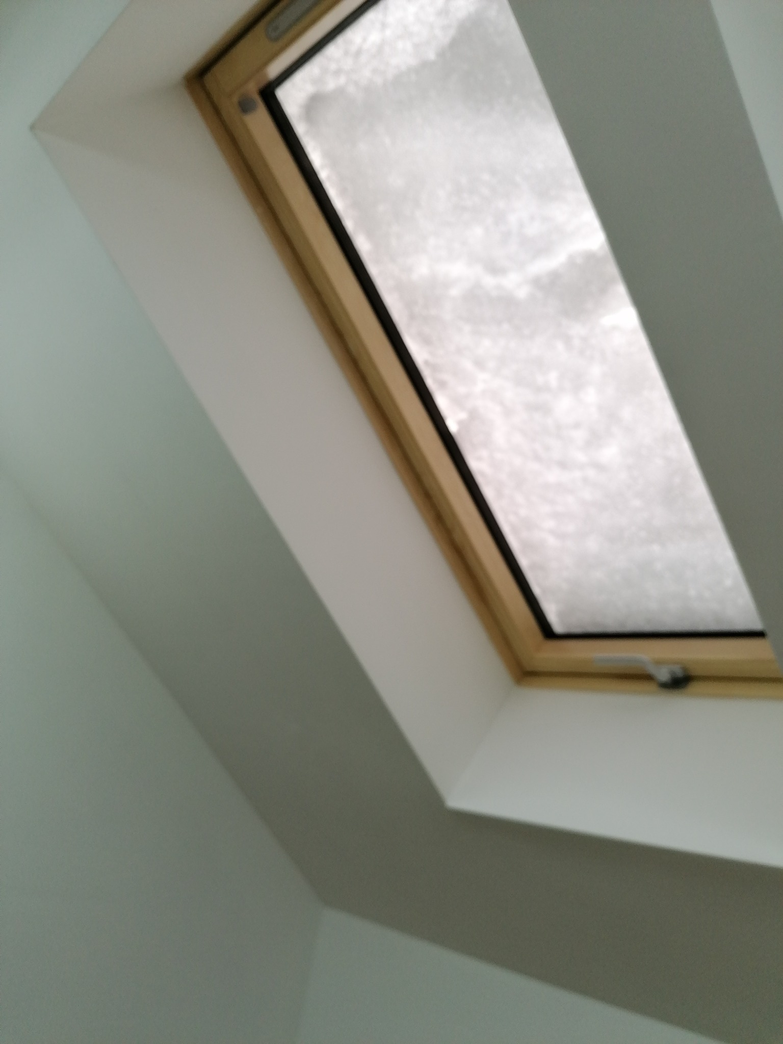 Okno dachowe z drewnianą ramą, widok z dołu na zamgloną szybę i szare ściany poddasza.