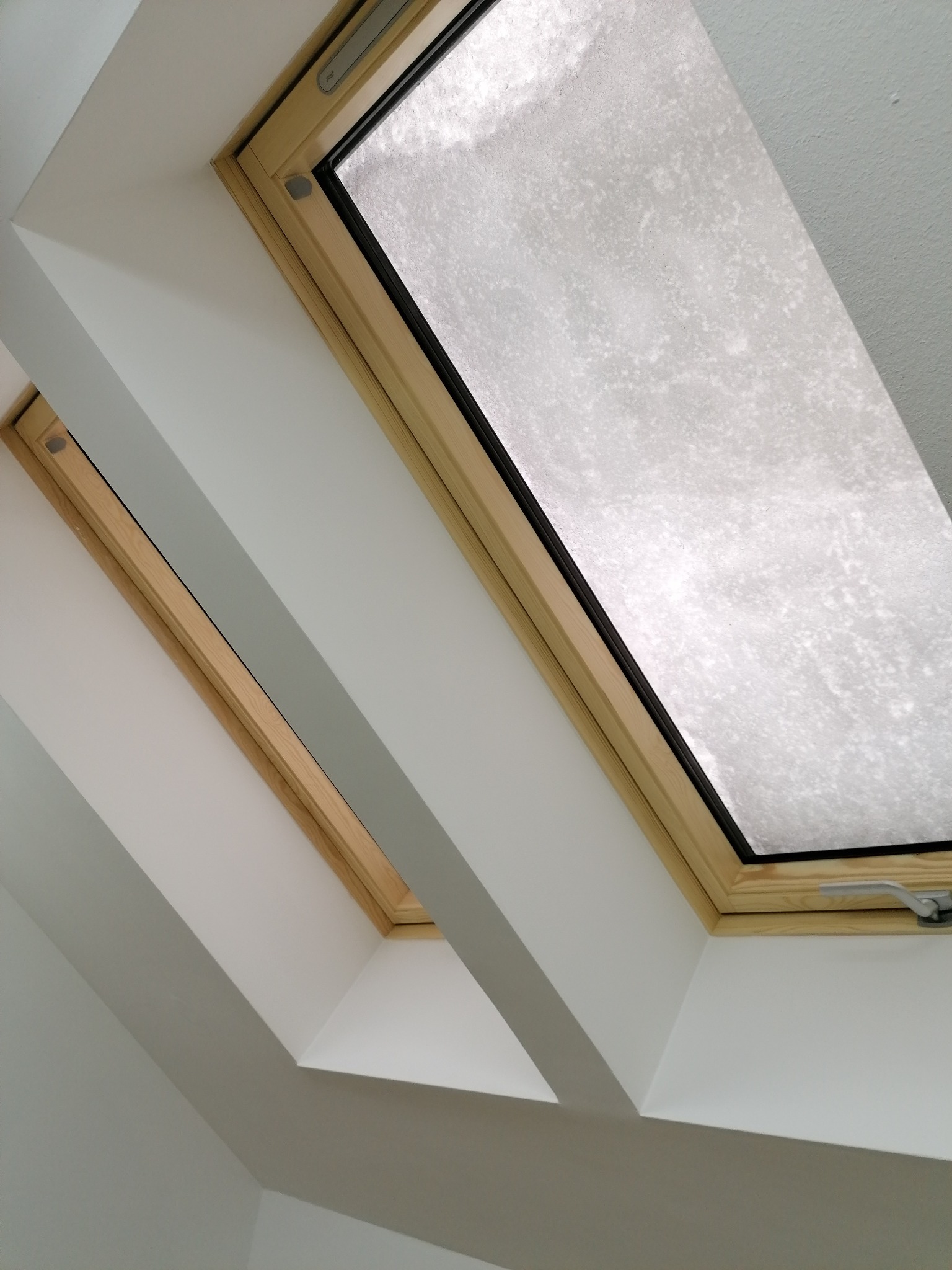 Dwa drewniane okna dachowe z widocznym oszronieniem na szybach, wkomponowane w białą ścianę pod skosem, uchwycone pod kątem.