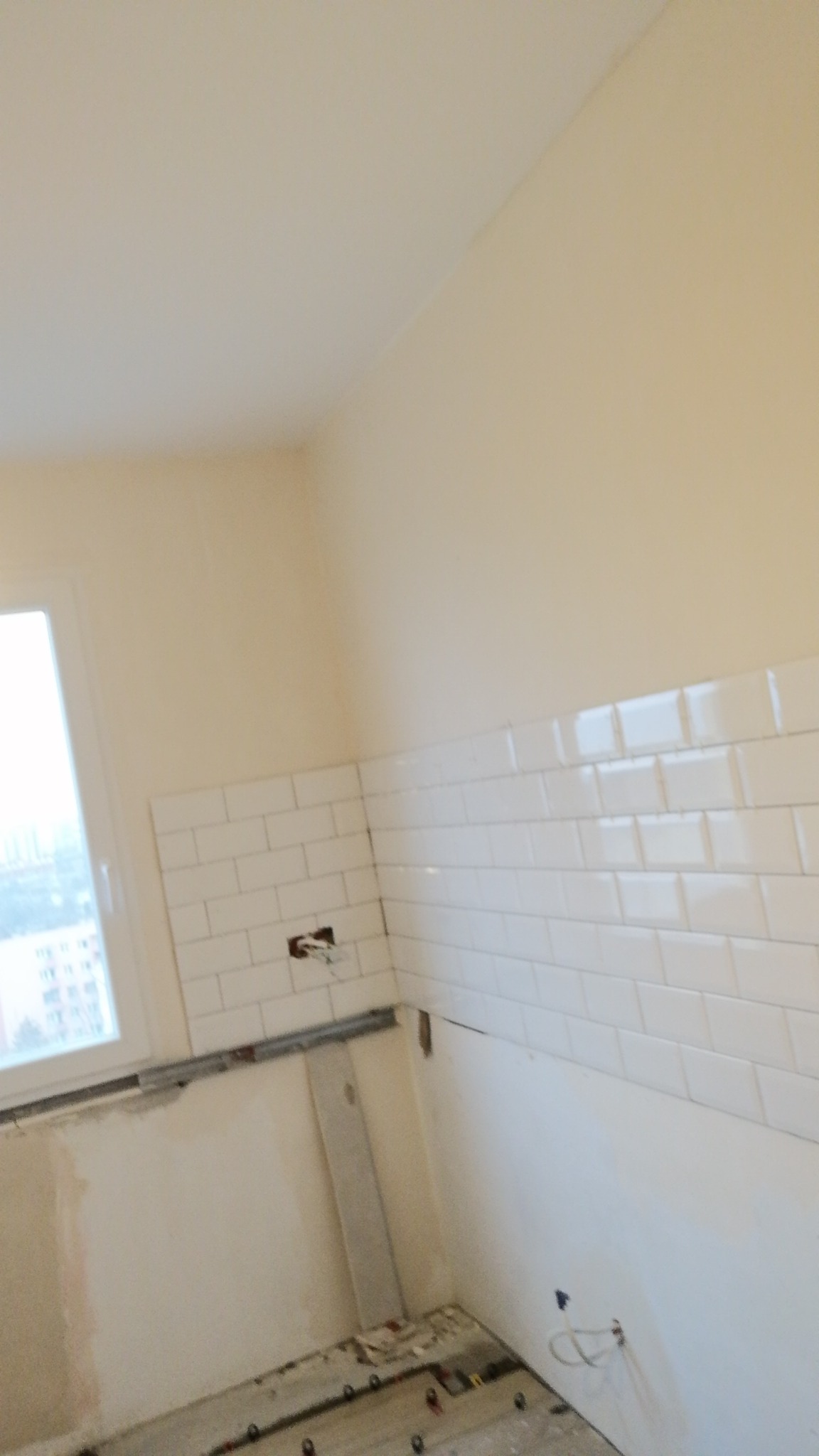 Ściana w trakcie układania białych, prostokątnych płytek, z widocznymi przewodami elektrycznymi i niedokończoną podłogą.