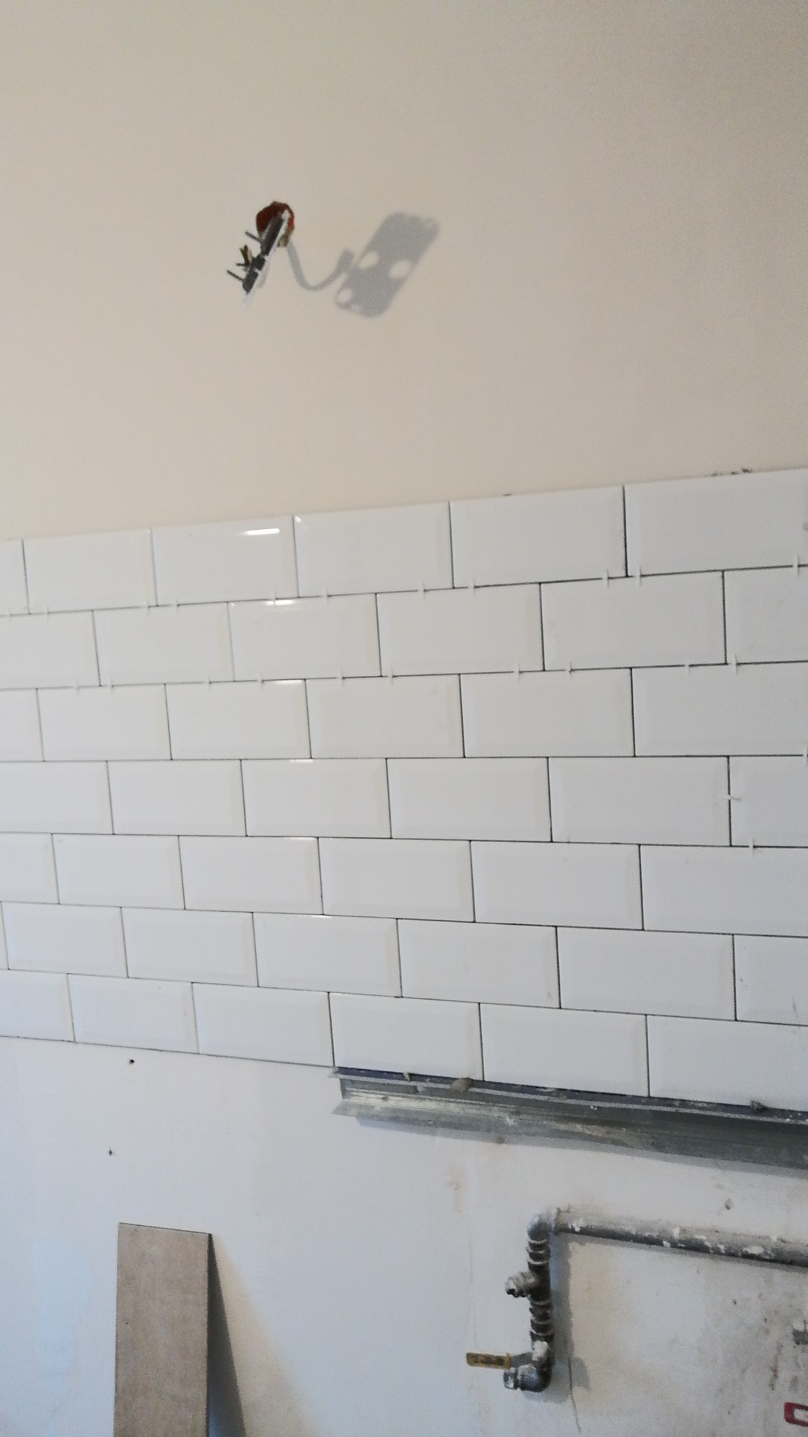 Ściana w trakcie układania białych, prostokątnych płytek, widoczne krzyżyki dystansowe, metalowa listwa poziomująca, instalacja elektryczna w suficie i fragment instalacji hydraulicznej przy podłodze.