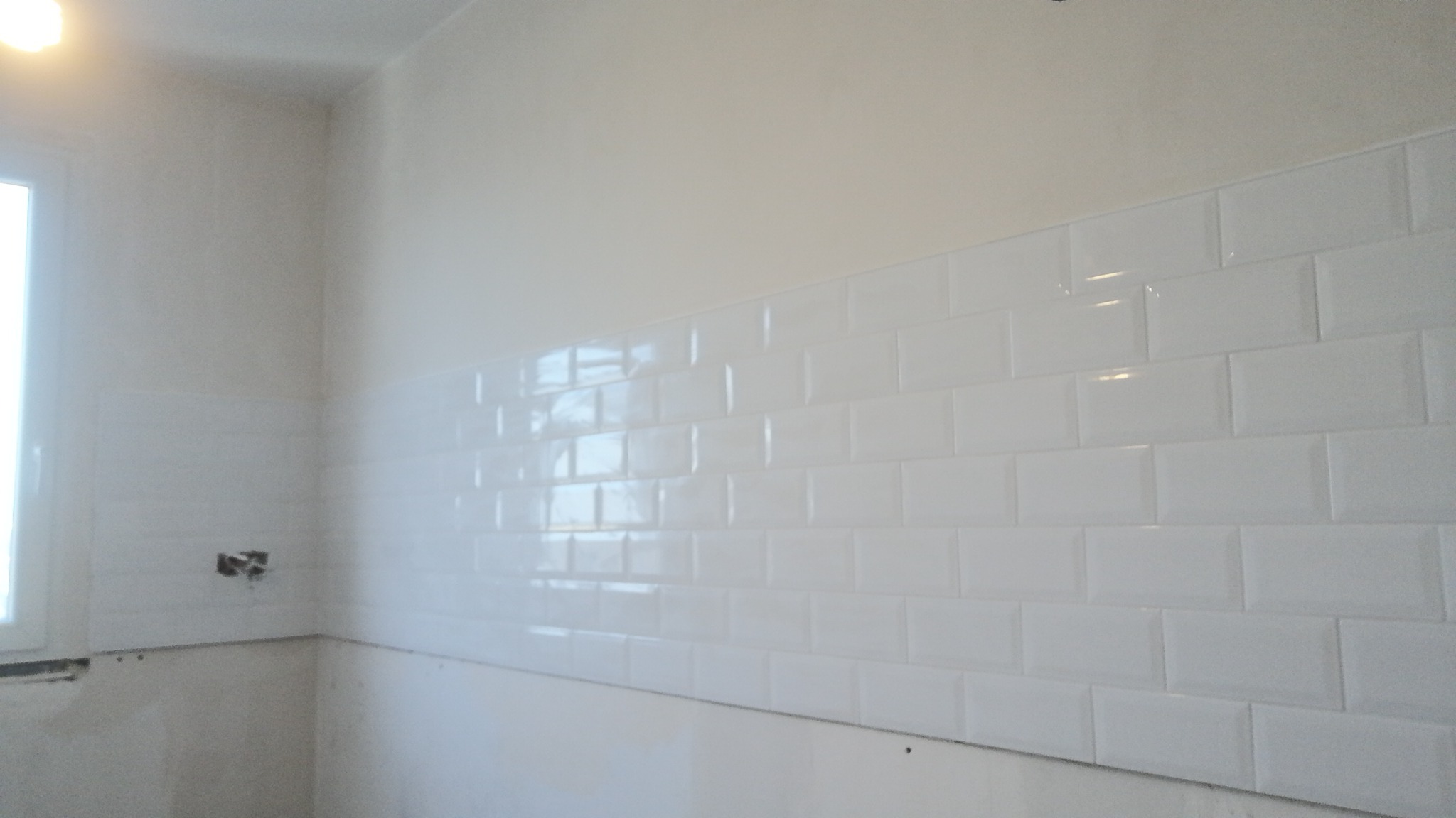 Ściana w trakcie wykańczania z białymi, błyszczącymi, prostokątnymi płytkami, widoczne niedokończone fragmenty przy oknie i puszka elektryczna bez osprzętu.