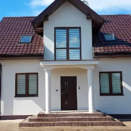 Nowoczesny dom jednorodzinny z brązowym dachem z blachodachówki, białymi ścianami elewacji i brązowymi oknami z roletami wewnętrznymi, widok z przodu na schody wejściowe.