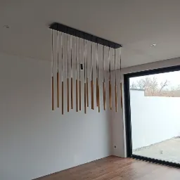 Nowoczesna lampa wisząca z podłużnymi, złotymi elementami na cienkich przewodach, zamontowana na czarnej, prostokątnej podsufitce, widoczna w przestronnym wnętrzu z dużym oknem tarasowym...