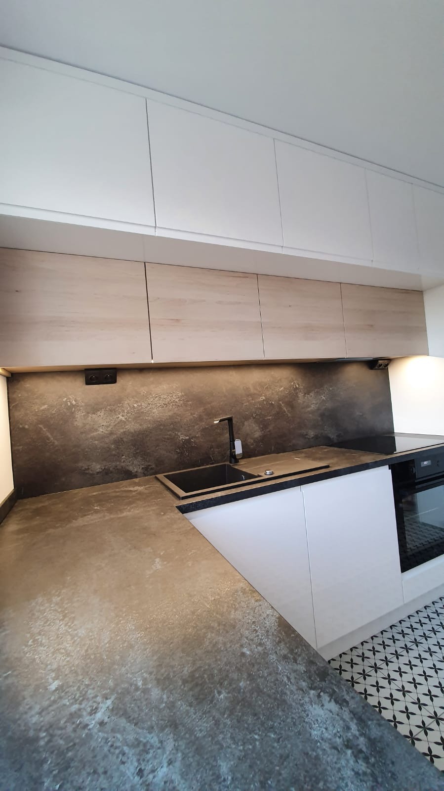 Nowoczesna kuchnia z drewnianymi szafkami wiszącymi, blatem imitującym beton, czarną armaturą i płytkami podłogowymi w geometryczny wzór.