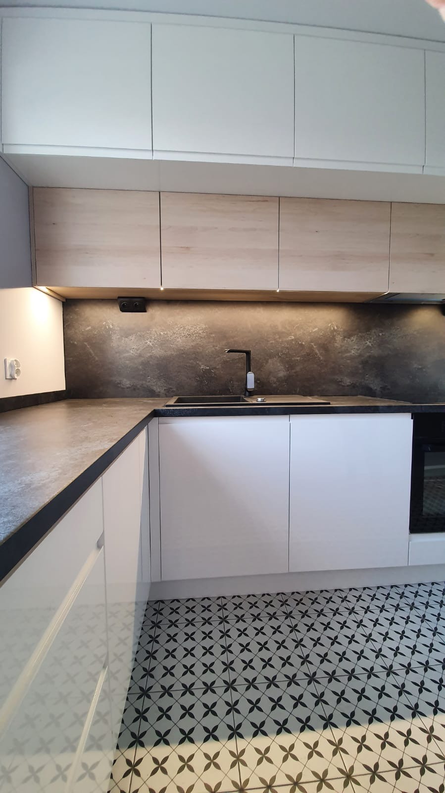 Nowoczesna kuchnia z białymi szafkami, drewnopodobnymi górnymi frontami, czarnym blatem i dekoracyjną mozaiką podłogową w stylu retro.