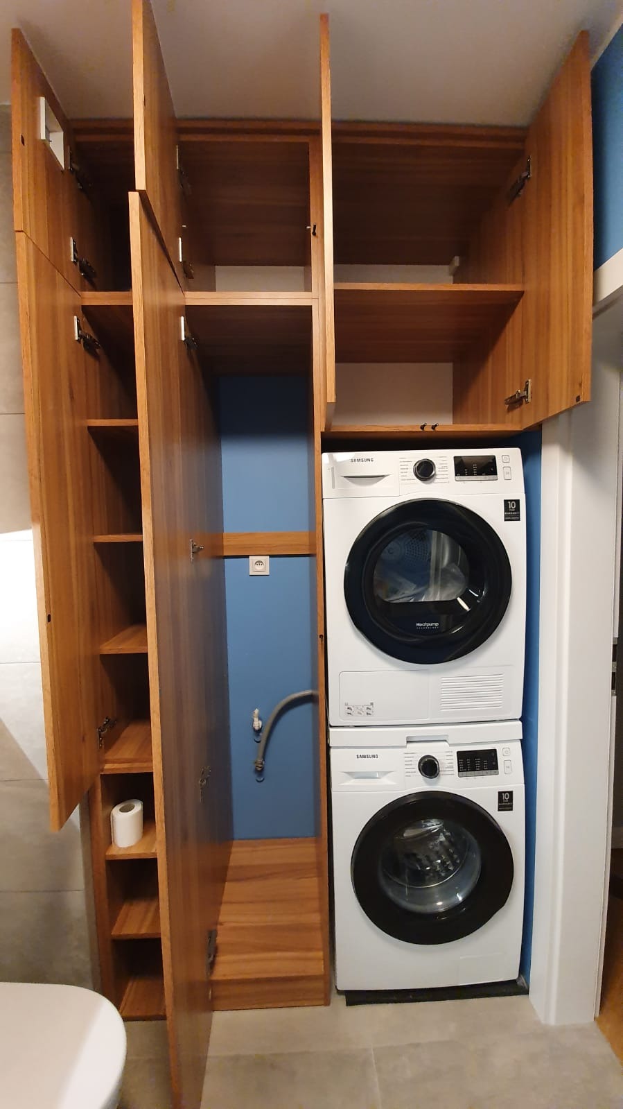 Wysoka, drewniana szafa z otwartymi drzwiczkami obok pralki i suszarki ustawionych jedna na drugiej, niebieska ściana w tle.