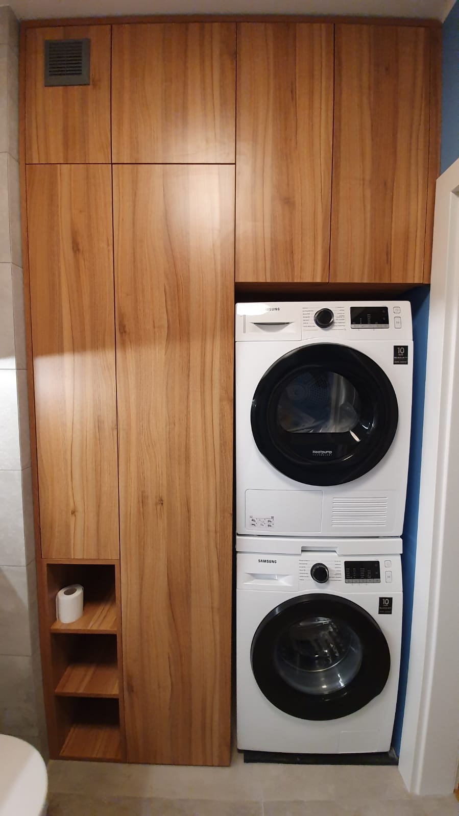 Wysoka zabudowa z drewnianymi frontami w odcieniach brązu, integrująca pralkę i suszarkę w łazience w Krzesinach Poznań, z otwartą półką na papier toaletowy.